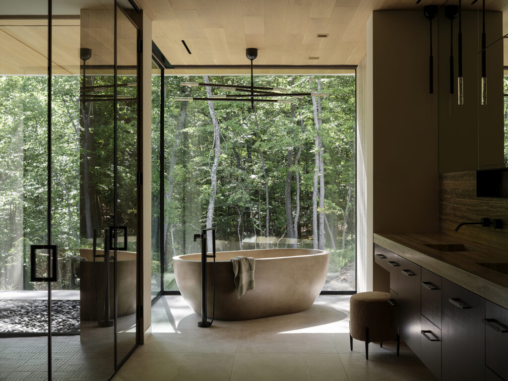 Biophilic design is showcased in this atrium style bathroom with floor to ceiling windows, wood paneled ceiling, large format travertine floors, and a modern abstract light fixture with a NativeStone concrete freestanding soaking tub as the focal point