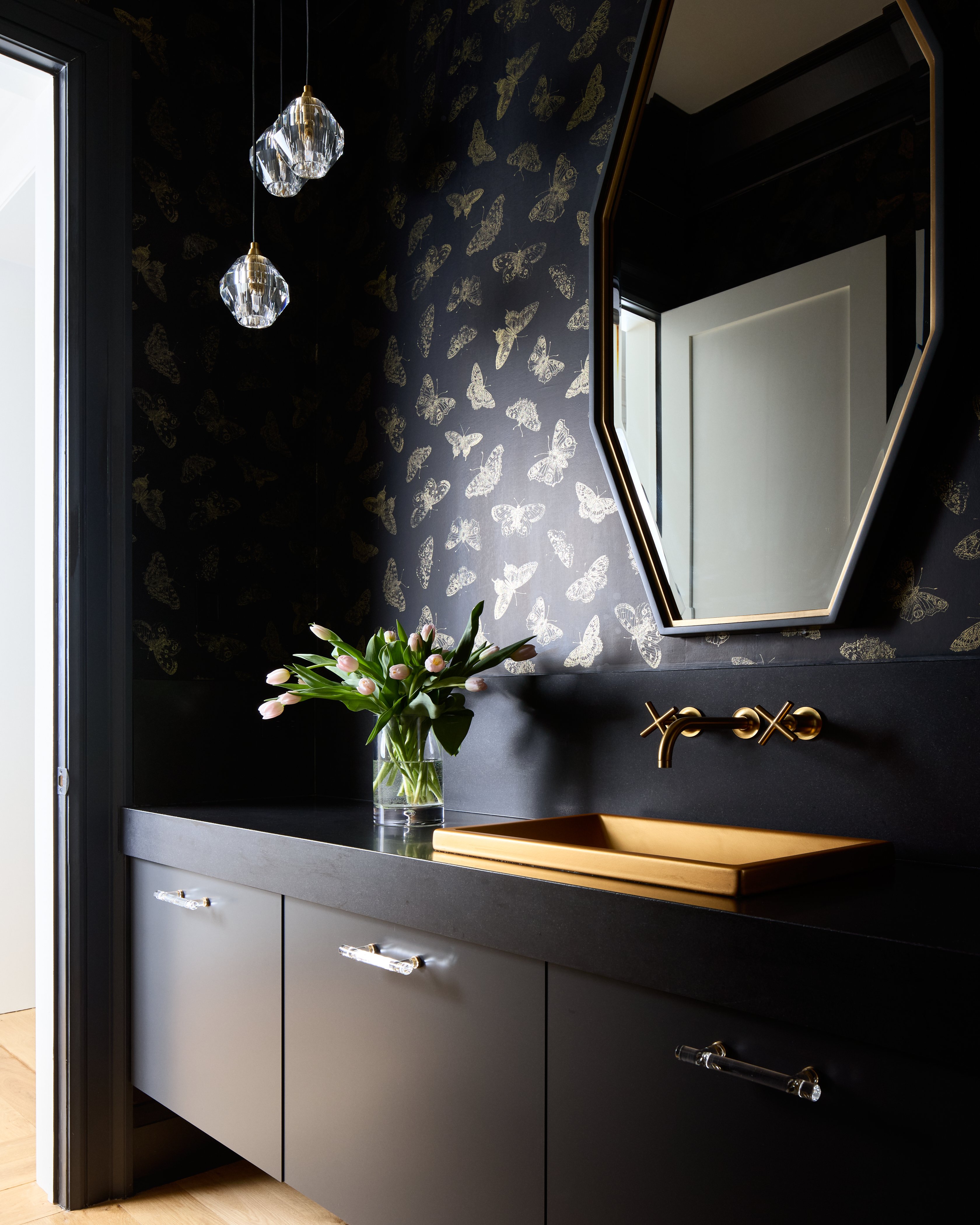 Moody powder room with black cabinets and a black stone countertop, butterfly wallpaper and a fireclay bathroom sink brushed in authentic 24k matte gold