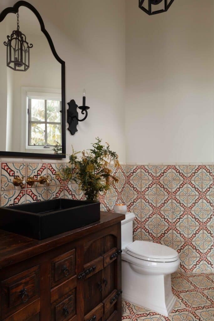 Bright, Spanish style tile bathroom featuring a handmade vanity and arched black bathroom mirror. Black Candelabras and a charcoal sink combine with a brass faucet.