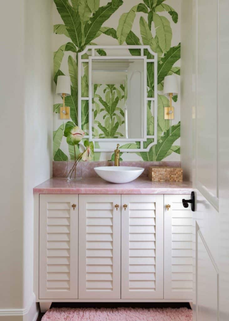 A Miami style bathroom features pink marble countertops and a vibrant wallpaper background featuring bright green plants. A white vessel sink sits on top of the counter.