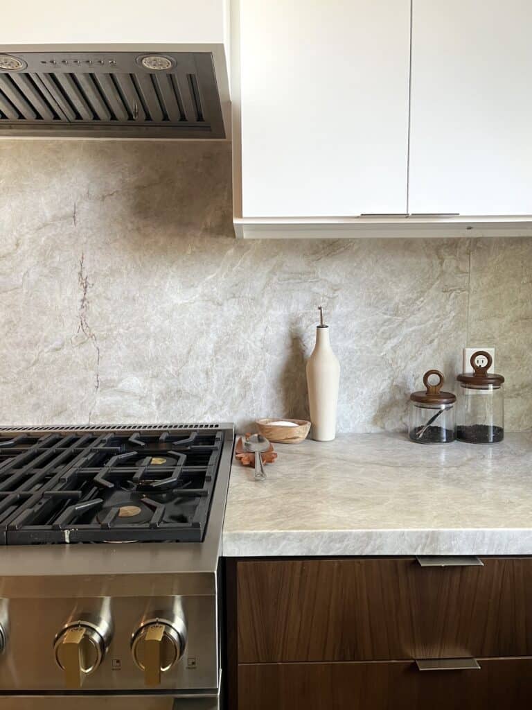 Close up of a kitchen stove with a marble countertop and marble backsplash. Showcasing organizational items such as a pinch pot for salt and other spice containers.