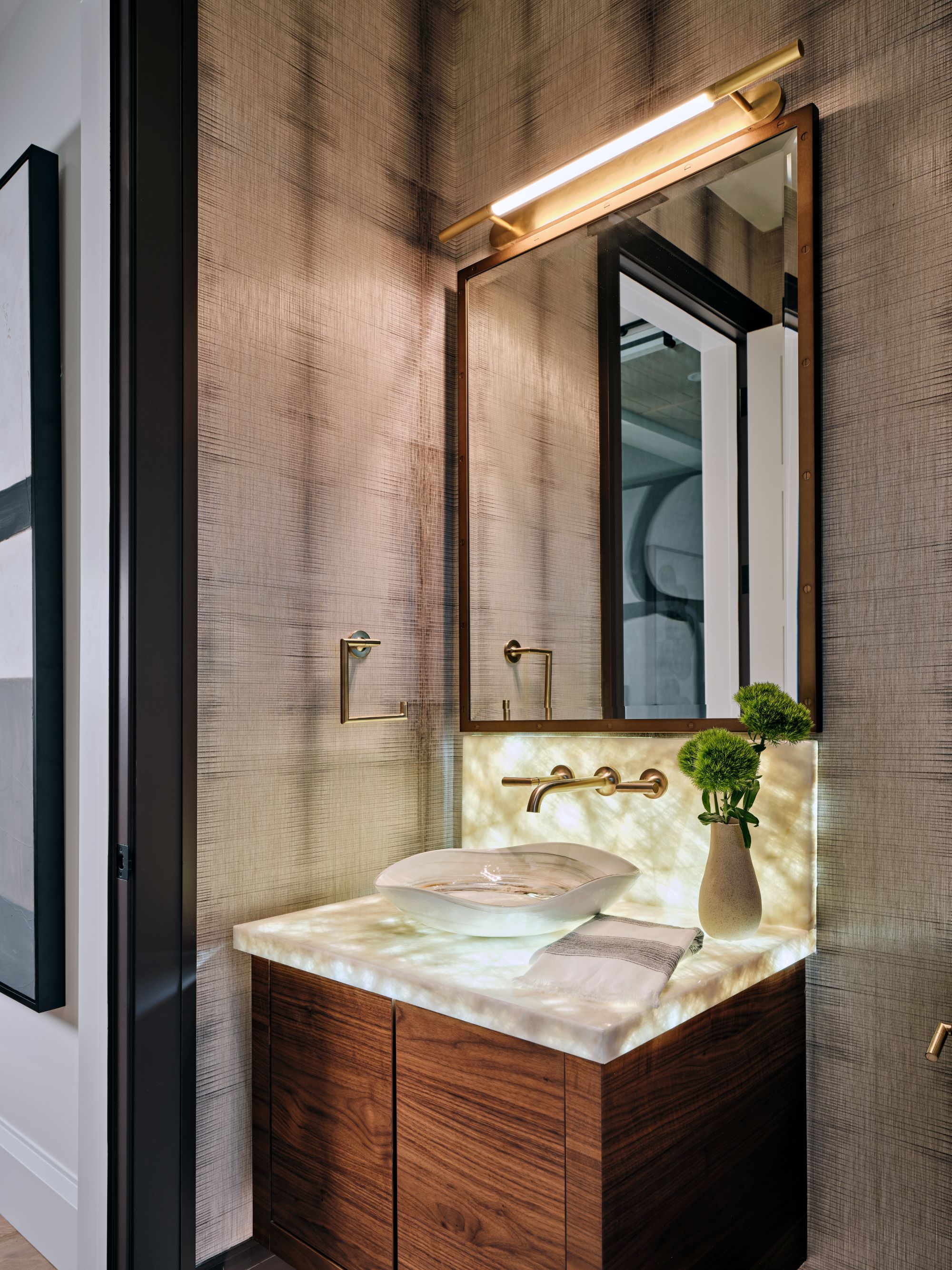 A Murano Glass vessel sink sits on top of a backlit marble countertop in a modern space featuring live walnut cabinets and silk wallpaper